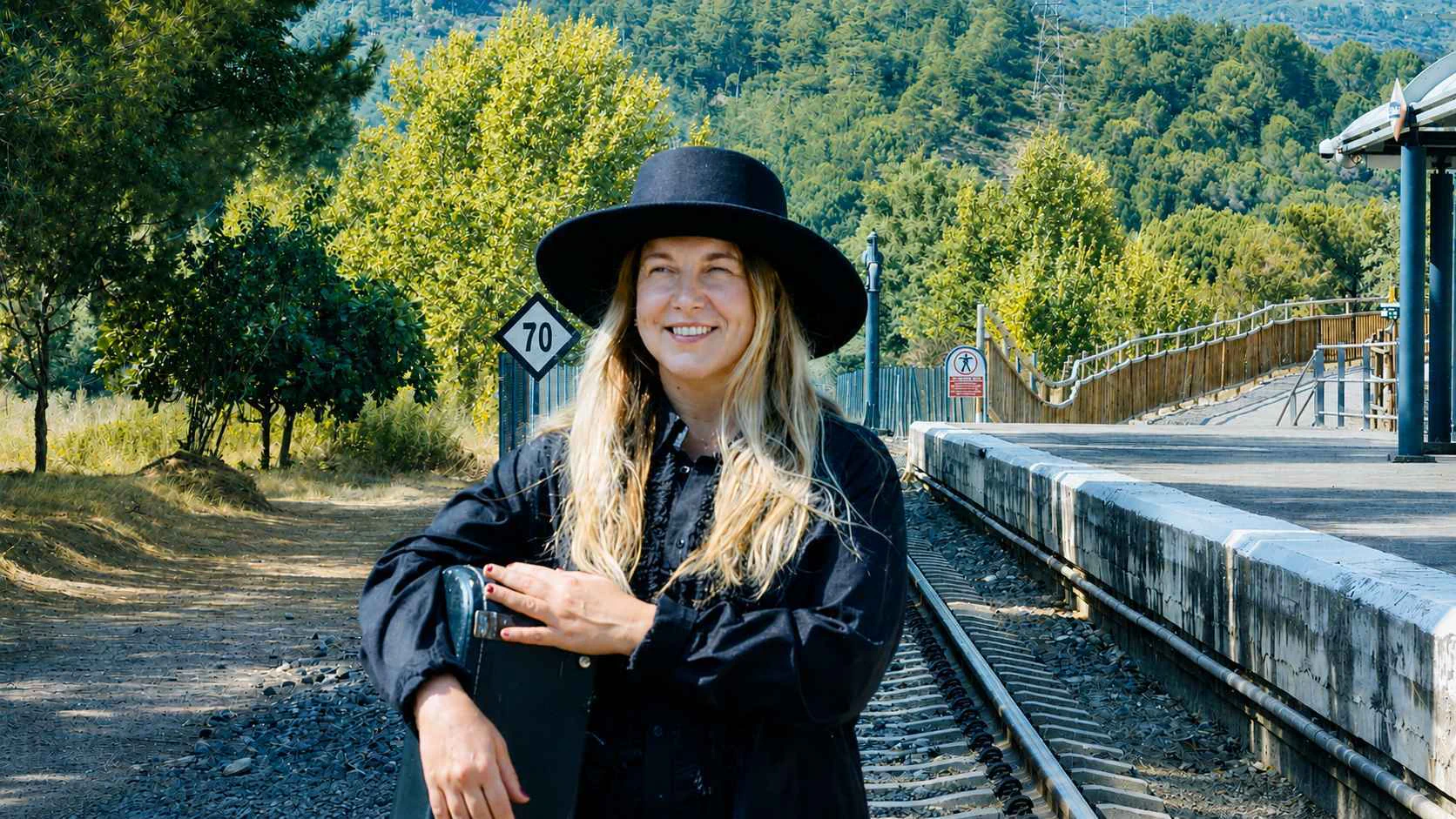 Victòria de Clascà at a rural train station, smiling in a black hat with an instrument case